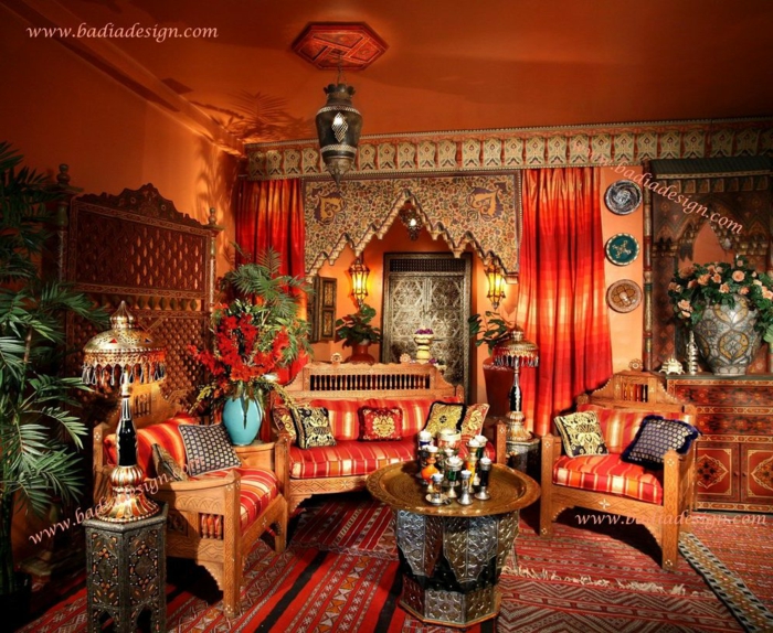reichlich dekoriertes Haus im marokkanischen Stil, eine Liege und zwei Sessel aus Holz, Kissen mit Streifenmuster, ein Kaffee- und ein Beistelltisch mit vielen Verzierungen, Designer-Stehlampe mit metallenem Schirm, vergoldetes Servierbrett in rznder Form, viele Blumen und Pflanzen, zwei Gardinen in Rottönen, Wand und Decke gestrichen in Orange, spanische Wand aus Holz, Teppiche mit unterschiedlichen Mustern, Kommode mit einer riesigen Vase darauf, drei dekorative Wandteller