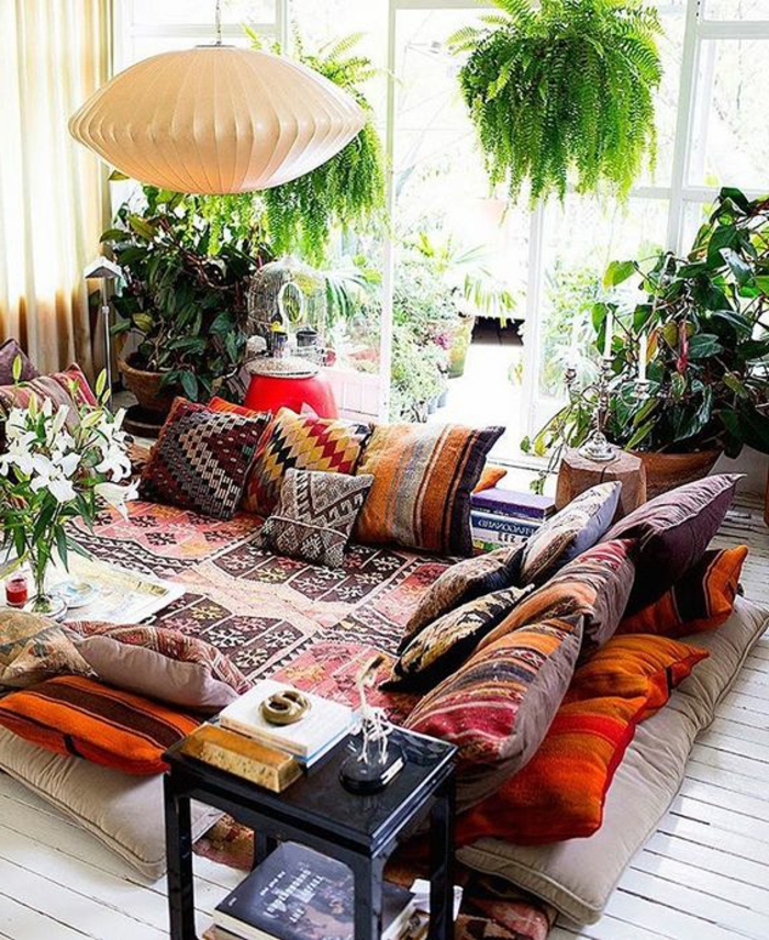weißer Holzboden mit kleinen Musterteppichen, marokkanische Kissen mit farbigen Mustern, viele grüne Pflanzen, zwei hängende Pflanzen, Kronleuchter aus weißem Papier, Relaxecke mit Terrassenaussicht