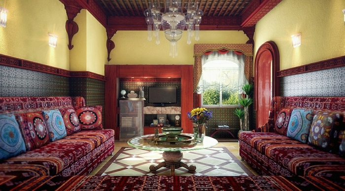 marokkanisches Wohhnzimmer mit flachem Fernseher in der Wandnische, gelbe Wände mit Mustertapeten in Blau, indirekte Wandbeleuchtung im Wohnzimmer, Kronleuchter aus Kristallen, runder Glastisch mit niedrigen Holzbeinen, Eckcouch mit marokkanischen Stoffen