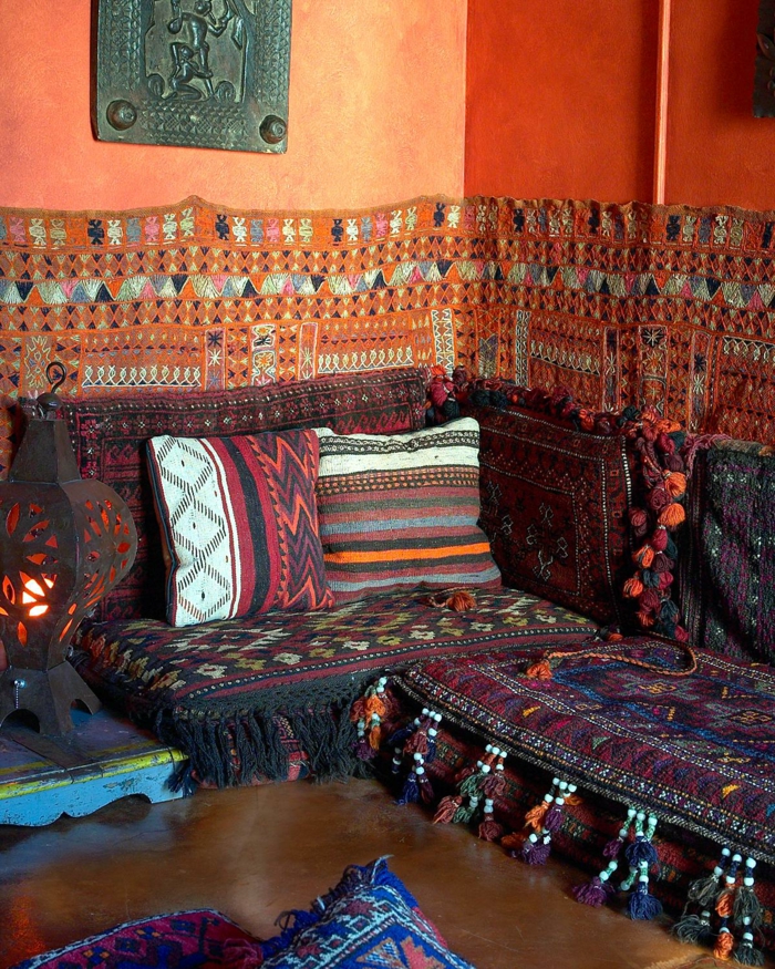 Couch in dunkle Farben, Couchdecke mit Troddeln, orange gestrichene Wand mit metallener Wanddeko, Stehleuchte aus Metall mit Schlitzerei