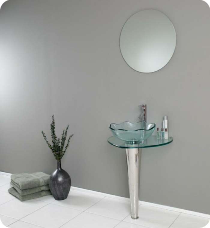 runder Wandspiegel über dem freistehenden Glasbecken, Bad mit grau gestrichener Wand und weißen Bodenfliesen, drei grüne Tücher neben einer grauen dekorativen Vase mit grünen Zweigen