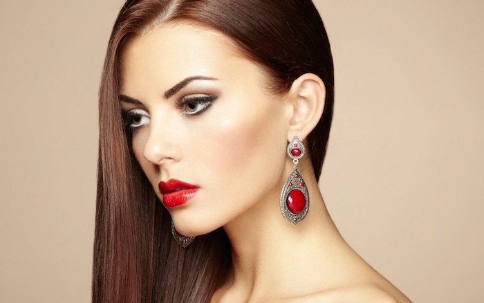 Lange glatte kastanienbraune Haare, braune Augen und Porzellanteint, roter Lippenstift, Ohrringe mit roten Steinen