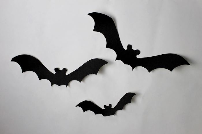 drei kleine schwarze fliegende fledermäuse aus papier, eine weiße wand mit einer halloween dekoration mit selbstgebastelten schwarzen fledermäusen mit schwarzen flügeln und ohren, eine fledermaus malen