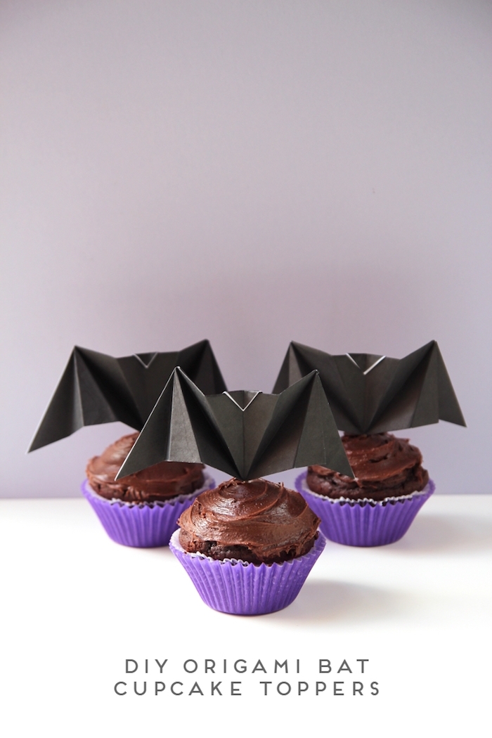 kuchen mit kleinen violetten muffinformen, drei kleine selbstgebastelte schwarze origami fledermäuse mit schwarzen flügeln aus papier, bastelideen für erwachsene