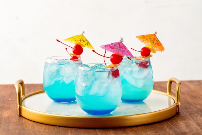 ein tisch aus holz und drei kleine gläser mit einer blauen limonade mit eis, kleinen roten kirschen und mit kleinen gelben, orangen und violetten cocktailschirmchen, eine erfrischungsgetränk selber machen 