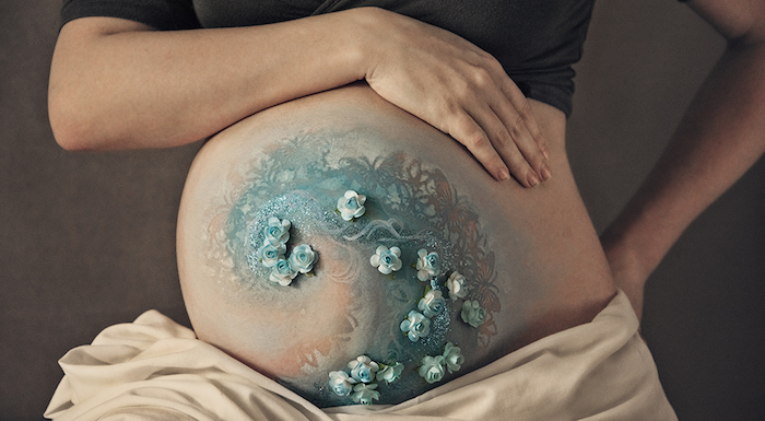 eine schwangere frau mit einem bild mit vielen kleinen blauen und weißen selbstgebastelten blumen aus papier, babybauch bemalen lassen