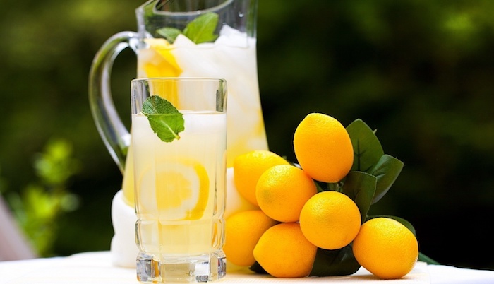 ein großer krug und ein glas mit vielen gelben zitronehn und mit eis und minze mit grünen blättern und einer gelben selbstgemachten limonade