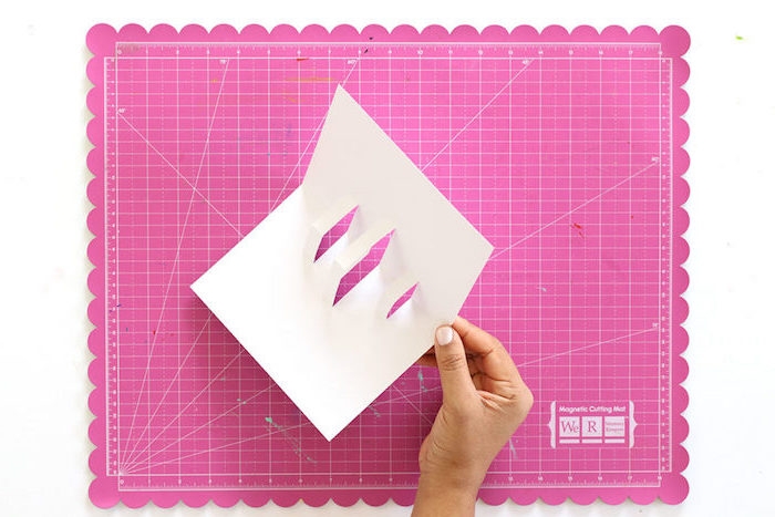 ein blatt papier, eine hand mit einem weißen nagellack, eine pop up karte selber machen, basteln mit papier, kleine weiße pop karte aus papier
