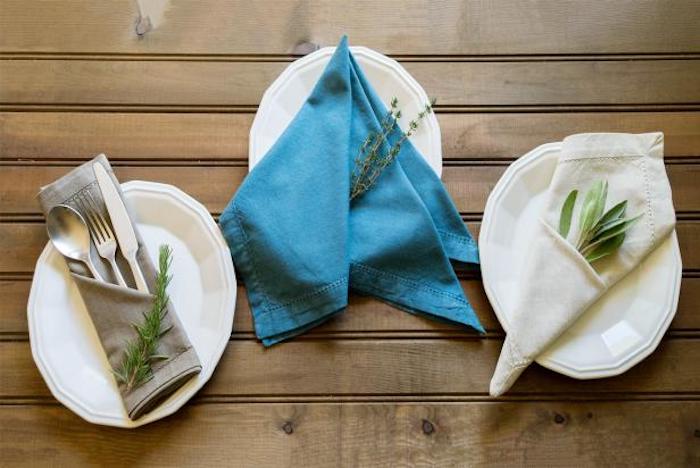 ein tisch aus holz, drei weiße teller mit grauen, blauen und weißen gefalteten servietten mit grünen pflanzen, eine graue bestecktasche mit einem löffel, einem messer und einer gabel