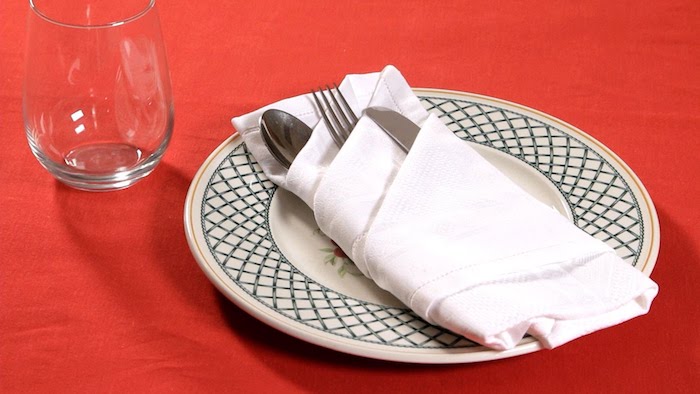 ein glas und ein weißer teller mit einer weißen bestecktasche mit einem löffel, einem messer und einer gabel, ein tisch mit einer roten decke, bestecktasche falten 