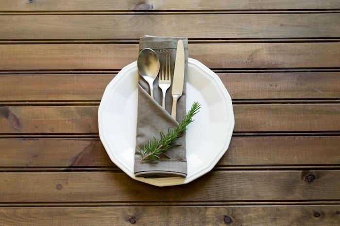 ein tisch aus holz mit einem weißen teller mit einer grauen serviette mit einem messer, einem löffel und einer gabel und mit grünen pflanzen, servietten falten einfach und wirkungsvoll