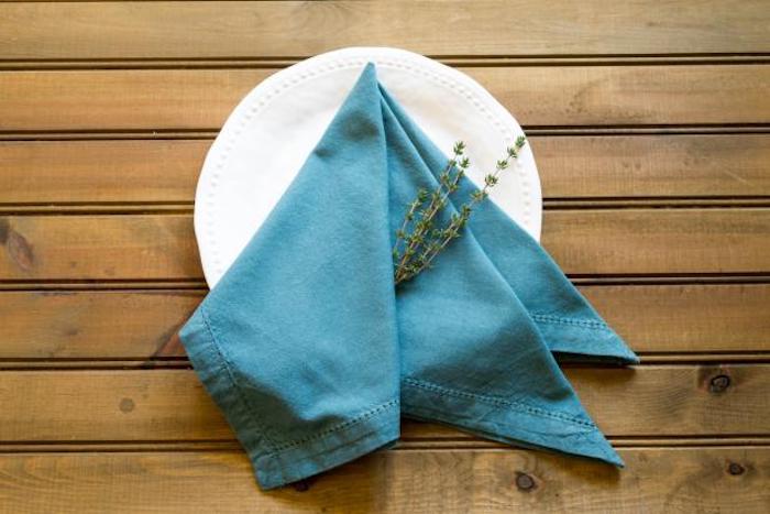 ein tisch aus holz, weißer teller mit einer großen blauen gefalteten serviette mit gr´nen pflanzen mit grünen blättern, servietten falten 