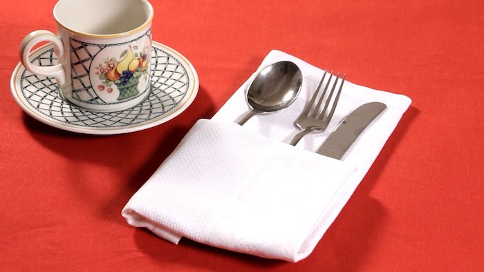 tisch mit einer roten decke, bestecktaschen falten, ein glas und eine weiße gefaltete bestecktasche mit einer gabel, einem löffel und einem messer