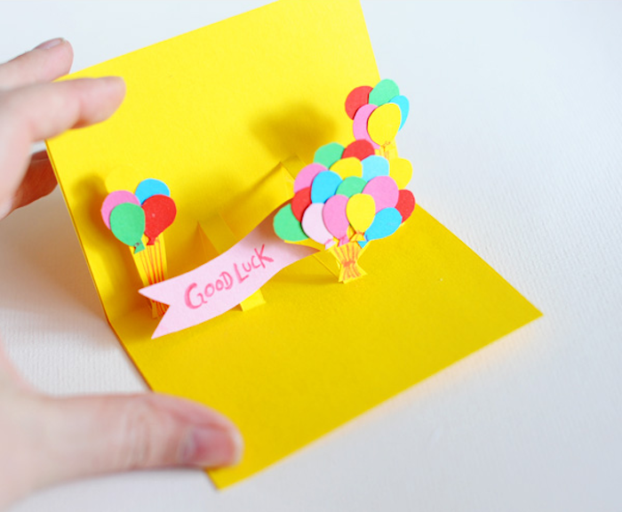 eine kleine gelbe pop up karte aus papier und mit vielen kleinen roten, violetten, grünen und gelben ballons, eine hand und eine karte zum geburtstag