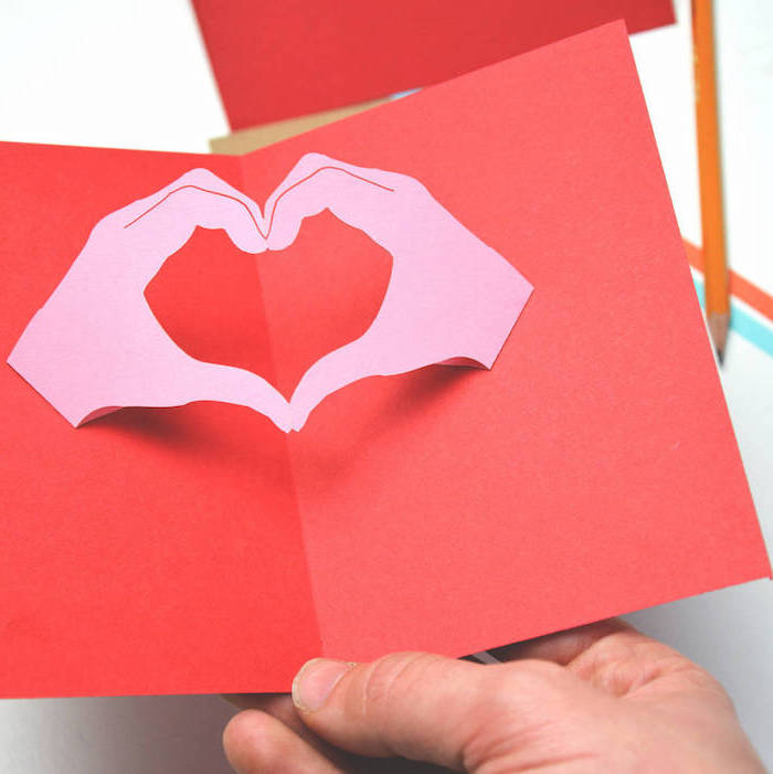 eine hand mit einer kleinen roten pop up karte mit zwei gebastelten violetten händen aus papier, ein kleiner oranger bleistift, pop uo karten vorlagen