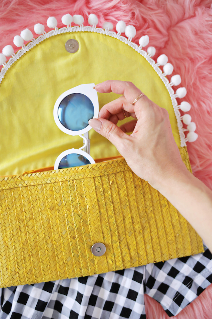 Gelbe Clutch mit weißen Pompons, Sonnenbrille darin, kariertes Hemd, rosa Decke