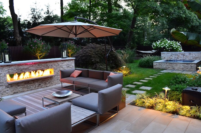 kleingarten gestalten, großer sonnenschirm, sofa mit sessel mit grauen kissen, feuerstelle aus naturstein