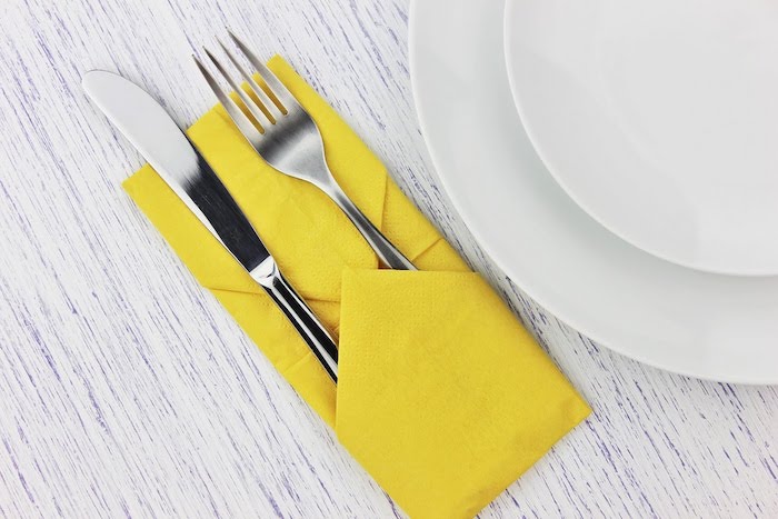 ein tisch mit einer großen gelben und gefalteten serviette und mit zwei weißen tellern, eine bestecktasche mit einem löffel und mit einem messer, ein weißer tisch aus holz, servietten falten hochzeit