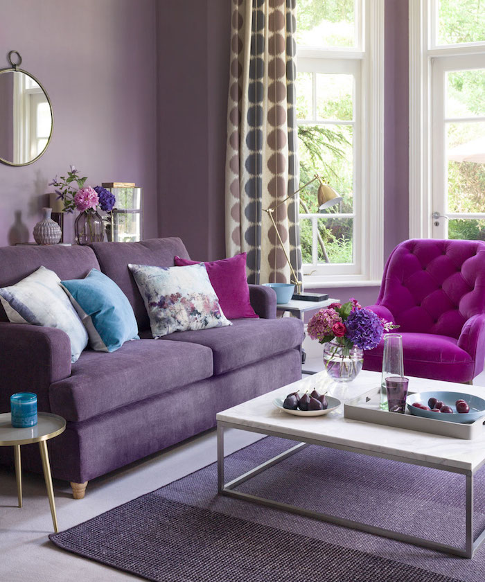 die farbe aubergine kombinieren, ein wohnzimmer gestalten mit einem kleinen weißen tisch und mit violetten sofas mit blauen, weißen und violetten kissen, eine vase mit pinken und violetten blumen, gardinen wohnzimmer, eine violette wand mit einem spiegel
