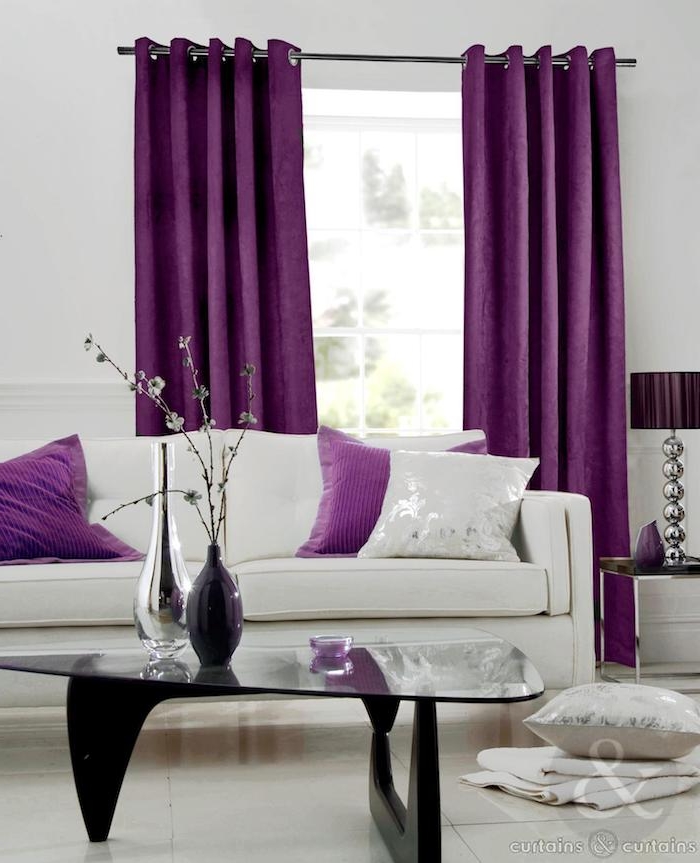 ein wohnzimmer mitr großen violetten gardinen und mit einem fenster, aubergine farbe kombinieren, ein kleiner schwarzer tisch und ein weißes sofa mit weißen und violetten kissen