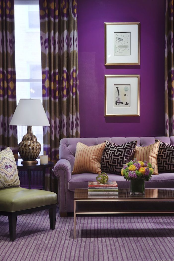 ein violetter teppich und ein kleiner tisch, ein fenster mit großen violetten gardinen, eine kleine weiße lamppe, orange und schwarze kissen, eine vase mit gelben und violetten blumen