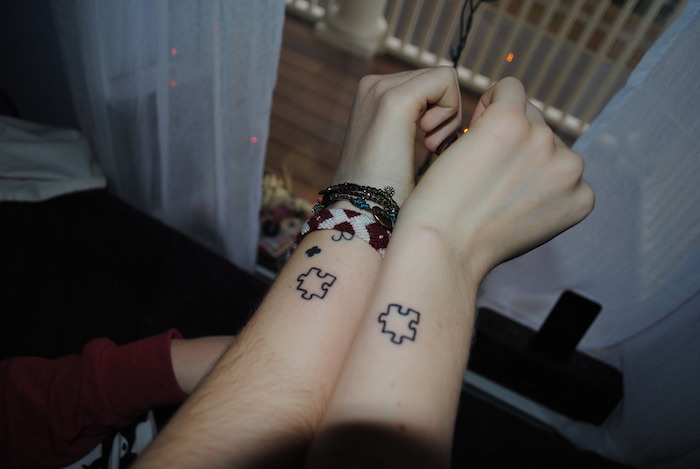 zwei hände mit armbändern und mit kleinen schwarzen puzzle tattoos, ein fenster und boden aus holz, liebes tattoo