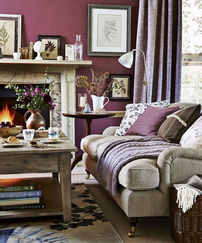 ein wohnzimmer mit einem kleinen tisch aus holz und einem sofa mit kleinen violetten und braunen und weißen kissen mit blumen, eine braune vase mit violetten blumen mit grünen blättern, aubergine farbe