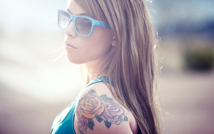 eine junge frau mkt einem blauen unterhemd und blaue brille, eine hand mit einem rosen tattoo mit zwei roten blumen und grünen blättern, tattoo frauen