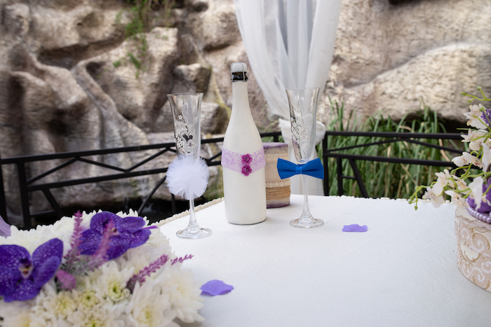 Tischdekoration für Hochzeit, weiße Weinflasche mit Spitze und kleinen Blüten dekoriert, Weingläser mit Fliege und Pompon