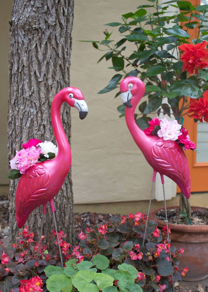 ein garten mit einem baum und mit zwei dekorativen pinken flamingos mit weißen und violetten blumen mit grünen blättern, flamongo deko, rote blumen und ein haus