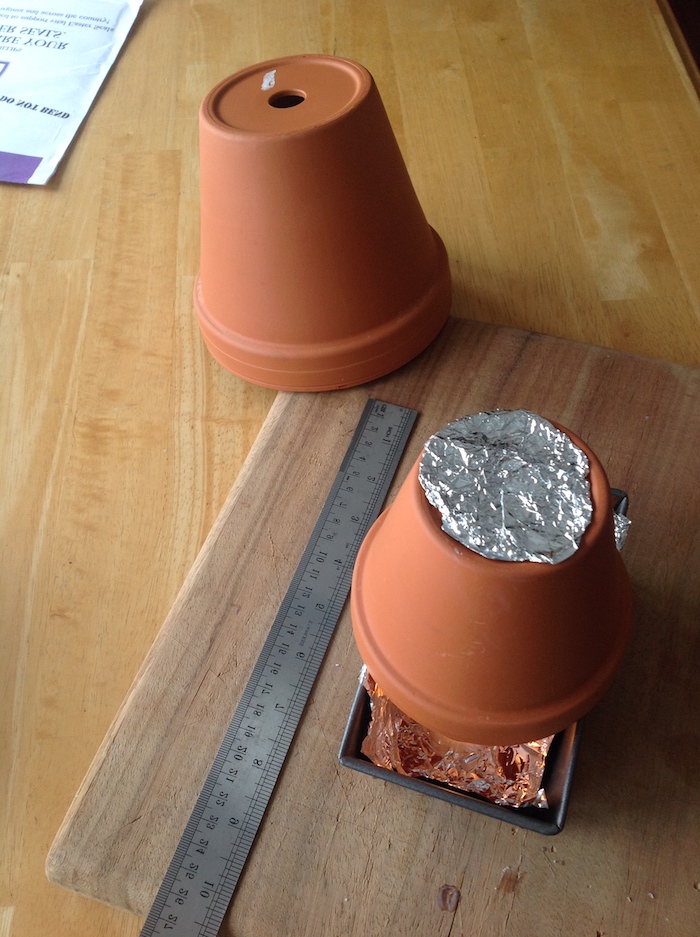 ein langes graues lineal und ein holzbrett, einen teelichtofen selber bauen aus zwei kleinen braunen blumentöpfen aus keramik, ein tisch aus holz