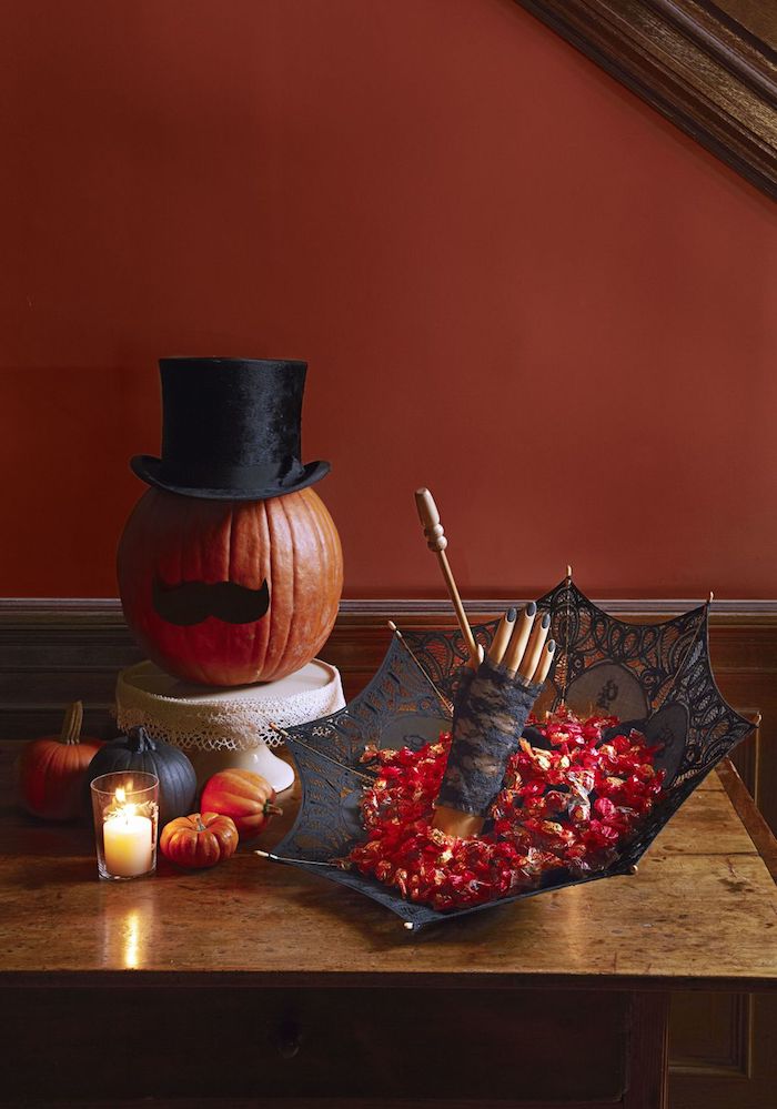ein schwarzer regenschirm und ein kleiner oranger halloween kürbis mit einem lustigen kürbisgesicht mit schnurrbart aus einem schwarzen papier und mit einem schwarzen hut, kleine kürbisse halloween und eine weiße kerze und ein tisch aus holz