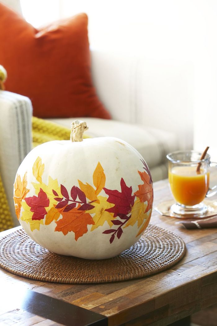 ein tisch aus holz, ein glas mit einem orangen saft, einen weißen halloween kürbis malen mit gelben, roten und orangen blättern, halloween dekoration selber machen
