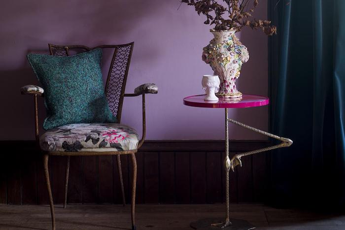 ein stuhl mit einem grünen kissen und ein kleiner violetter flamingo tisch mit einer bunten vase mit pflanzen und eine vilette wand