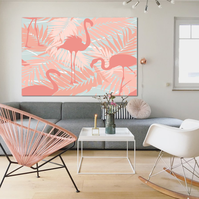 ein graues sofa und ein bild mit vielen pinken flamingos mit pinken federn und mit pinken und blauen palmen, ein kleiner tisch mit einer kerze und einer vase, boden aus holz und zwei stühle