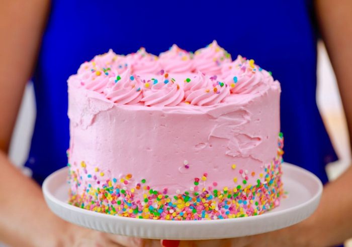 rosa Torte mit bunten Streuseln, eine Frau mit blauem Kleid trägt sie, Geburtstagstorte selber machen