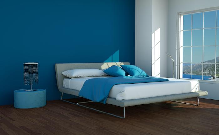 eine weiße wand mit fenster in einem schlafzimmer mit einem bett mit weißen und blauen kissen und mit einem braunen boden aus holz und einer blauen wand