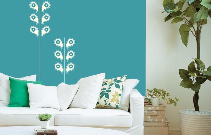 große blaue wand mit weißen blumen, ein wohnzimmer mit einem weißen sofa mit weießn und grünen kissen mit blumen, ein topf mit einer großen grünen pflanze 
