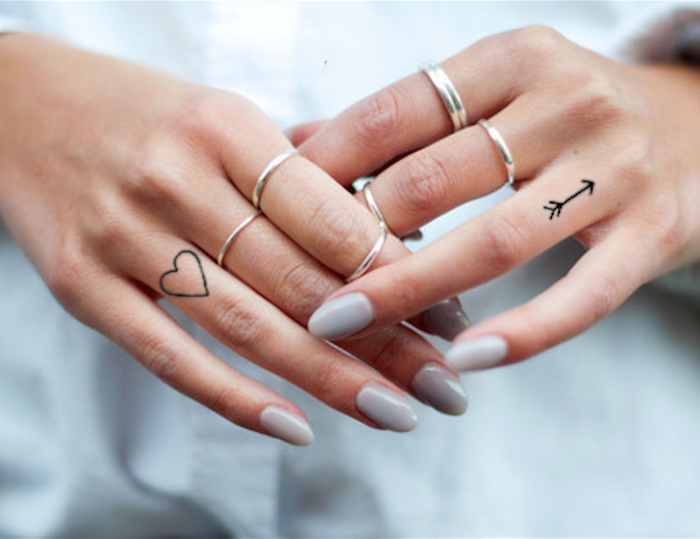 Zwei kleine Finger Tattoos, Herz und Pfeil, silberne Ringe, weißer Nagellack
