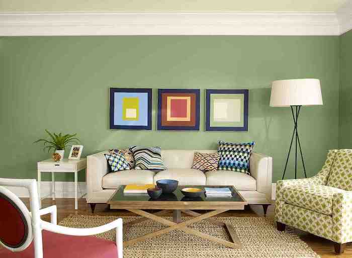 große grüne wand mit drei bildern mit blauen bilderrahmen und eine große weiße lampe, ein kleiner brauner tisch aus holz, ein sofa mit kissen, wohnzimmer streichen ideen