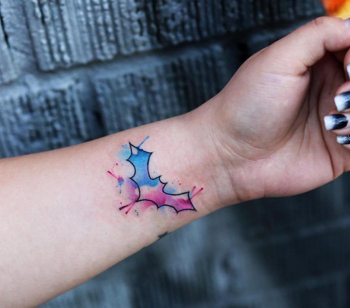 eine hand mit einem kleinen batman tattoo aquarell mit einer fledermaus mit blauen und violetten flügeln, tattoo am handgelenk, frauen tattoos