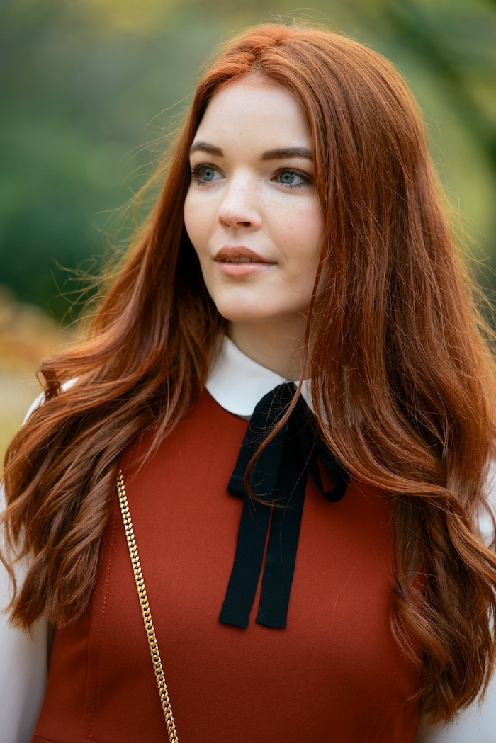 Lange kupferfarbene Haare mit Mittelscheitel, kühler Hautteint und blaue Augen, rotes Kleid mit weißem Kragen und schwarzem Band
