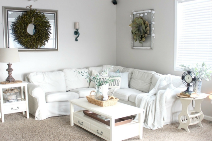 weiße landhausmöbel wohnzimmer, großer blumenkranz in grün hängt an der wand als deko, weißer tisch großes ecksofa