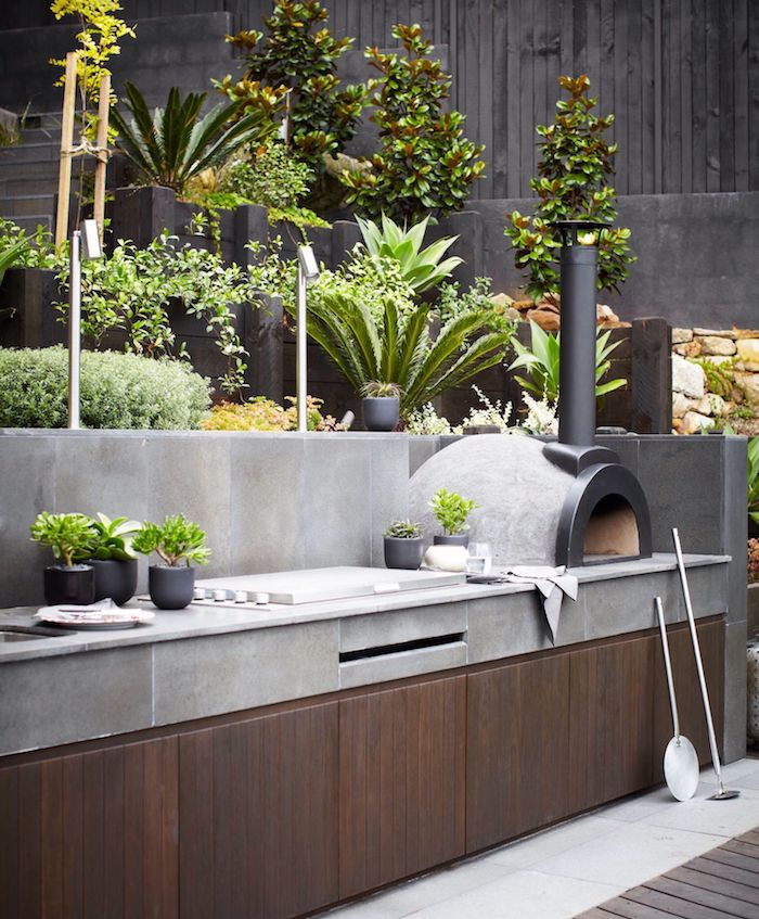 eine graue outdoor küche mit einem weißen waschbecken, ein garten mit grünen pflanzen, ein grill und kleine graue blumentöpfe 