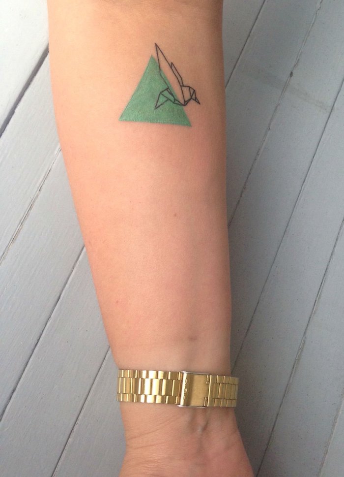 ein weißer tisch aus holz und eine hand mit einer goldenen uhr, eine hand mit einem aquarell tattoo mit einem grünen dreieck und mit einem fliegenden vogel