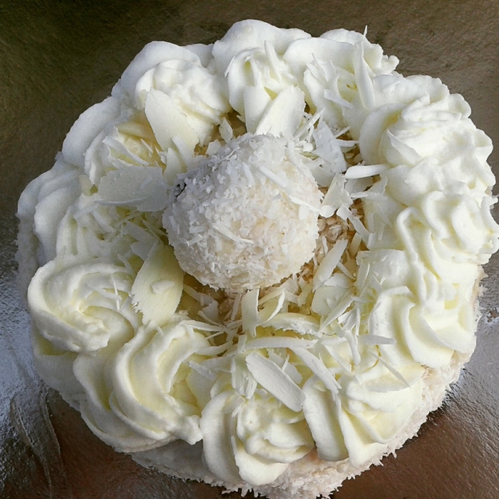 eine kleine süße Torte mit Raffaello Praline in der Mitte, Kokosraspeln, weiße Glasur