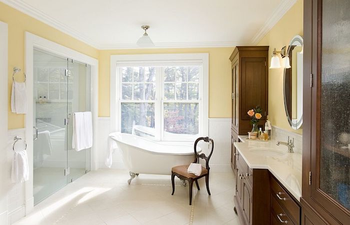 weiße kleine freistehende badewanne im badezimmer mit gelben wänden und großen weißen fenstern, ein brauner stuhl aus holz, badezimmer spiegelschrank