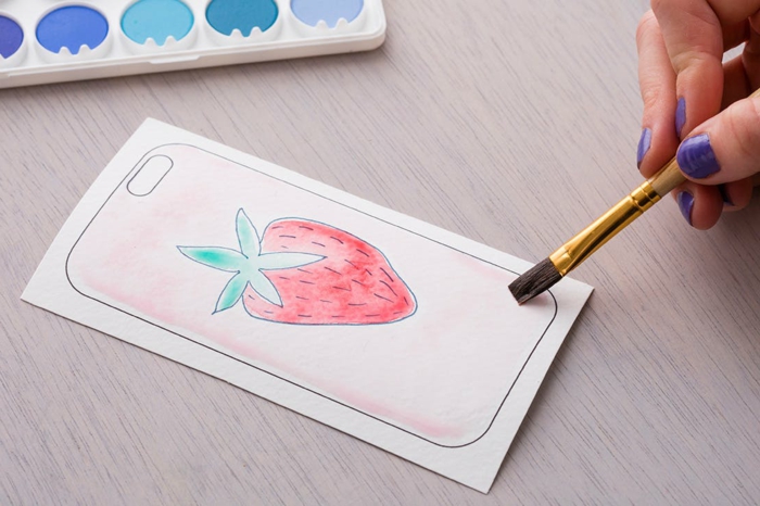 DIY Anleitung für Handyhülle selbst designen, Zeichnung von Erdbeere bemalt in pink und grün, Hand mit lila Nagellack