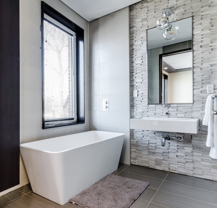 weiße freistehende badewanne im badezoomer mit einem fenster und spiegel, boden aus grauen badezimmer fliesen