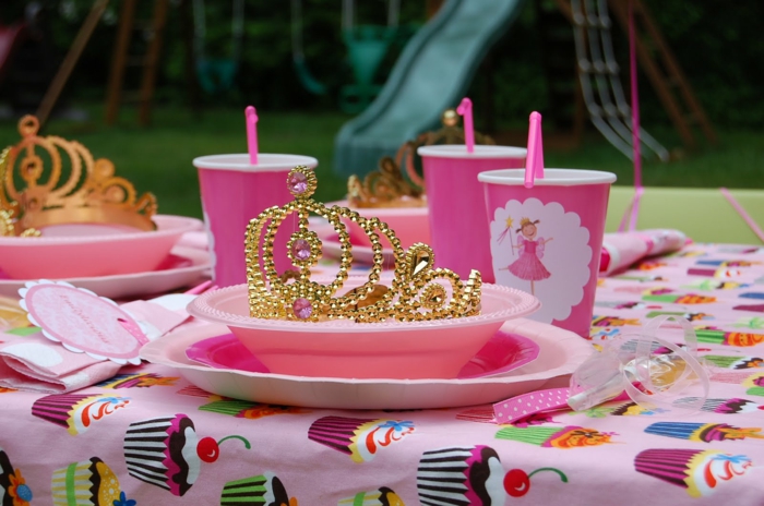ein Kindergeburtstag, rosa Becher mit Prinzessinnen, eine goldene Krone, bunte Tischdecke mit Süßigkeiten versehen, Geburtstagsdeko